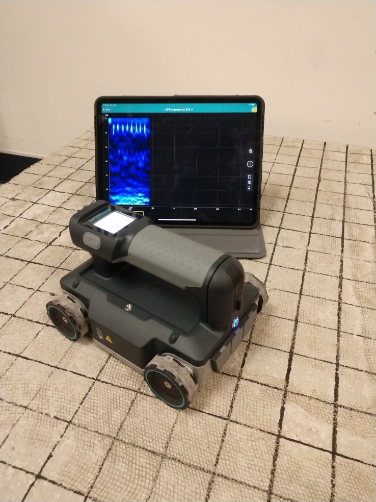"Figure 5. Proceq Live Wireless GPR antenna with range of 0.9 to 3.5 GHz. This figure shows the wireless GPR system with the tablet in the back showing a B-scan of the data and the manual antenna cart in the front of the photo."