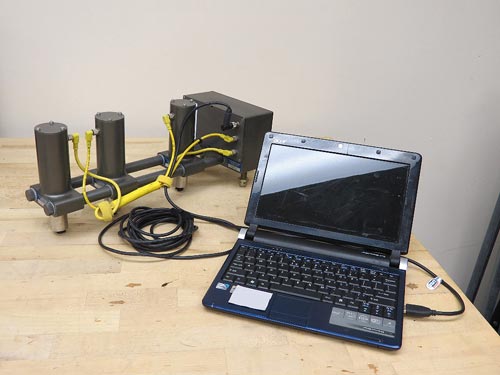 "USW Testing System.  This photo shows ultrasonic surface wave testing equipment, which has three vertical, cylindrical objects and one metal box, all attached to two large, rounded, horizontal connecting bars. The two cylindrical objects closest to the box are sensors. The third cylindrical object on the right end of the connecting bar is an impact source. Yellow cables run from the metal box to each of the cylindrical objects. A laptop collect all the data with a cable attached to the tool."